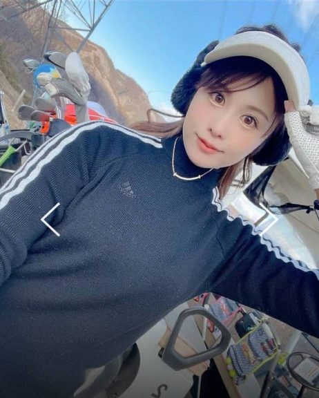 黒いadidasのゴルフウェアを着て、白いキャップと黒いいやーマフラーを付けた色白の女性がこちらを見ている。後ろには冬の山々が見える。